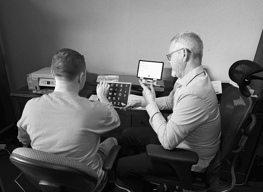 Sam using a letter board to communicate with J Brad, demonstrating spelling to communicate and presuming competence.